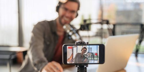 Male trendy podcast creator recording video podcast on smartphone. Selective focus on smartphone camera screen with man podcaster vlogger in headphones and glasses shooting live video for his channel